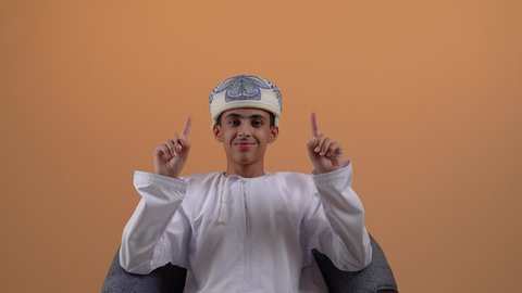 تحديد موقع معين، تصوير مقرب لولد مراهق عربي خليجي عماني يرتدي الدشداشة والعمامة يجلس على الكرسي يشير بأصبع السبابة لأعلى الى هدف محدد، مفهوم النقر والضغط والتفاعل مع العناصر الهولوجرامية، خلفية بيج