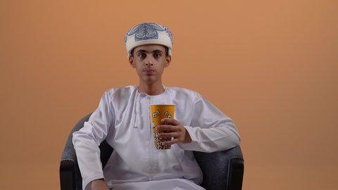 انشطة ترفيهية منزلية، الاستمتاع بتناول الفشار، بورتريه لولد مراهق عربي خليجي عماني يرتدي الدشداشة والعمامة يجلس على كرسي مريح يحمل بيده علبة الذرة، سناكات ومقرمشات لذيذة، خلفيه بيج