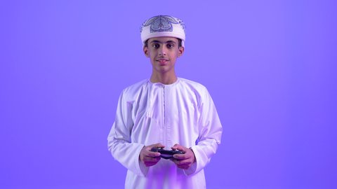 تعابير التركيز والإندماج ، ألعاب الفيديو الترفيهية، تصوير مقرب لولد مراهق عربي عماني خليجي يرتدى الدشداشة والعمامة يحمل بيده عصا التحكم عن بعد بايماءات السعادة و الحماس، تقنيات وألعاب حديثة، قضاء أوقات ممتعة ، خلفية زرقاء
