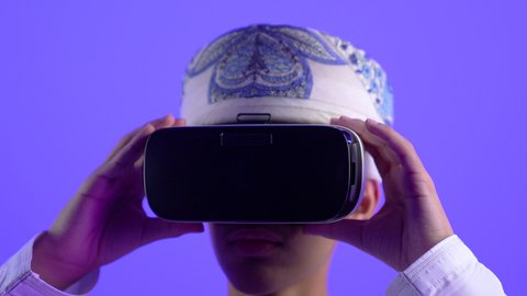 المتعة والتسلية بالمشاهدة عبر نظارة الواقع الافتراضي، محاكاة العالم الخيالي، تصوير مقرب لولد مراهق عربي خليجي عماني يرتدي الدشداشه والعمامة يضع نظارات ثلاثية الأبعاد (الفي ار) بإيماءات السرور, استخدام جهاز تقني حديث ومتطور، خلفية زرقاء