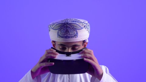 محاكاة العالم الخيالي، المتعة والتسلية بالمشاهدة عبر نظارة الواقع الافتراضي، تصوير مقرب لولد مراهق عربي خليجي عماني يرتدي الدشداشه والعمامة يضع نظارات ثلاثية الأبعاد (الفي ار) بإيماءات السرور, استخدام جهاز تقني حديث ومتطور، خلفية زرقاء
