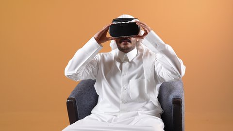 الاندماج والتفاعل ومحاكاة العالم الخيالي، تصوير لرجل عربي خليجي قطري يرتدي الثوب التقليدي والشماغ ونظارات الواقع الافتراضي يجلس على كرسي مريح يقوم بمد يديه الى الامام بتعابير صد شيء ما والدفاع عن النفس، شاب سعودي يستخدم نظارة الفي آر