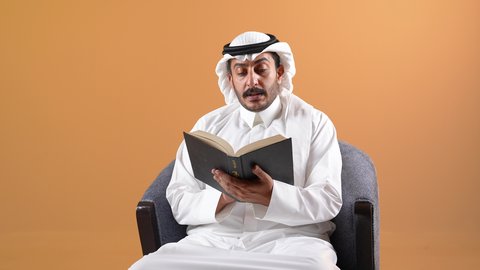 Developing skills, expanding knowledge and ideas, increasing intellectual stock, spending free time usefully, enjoying reading books, stories and novels, a picture of a young Qatari Gulf Arab man wearing traditional thobe and shemagh sitting on a chair and holding a book in his hands