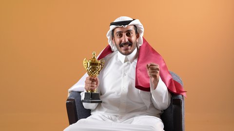 تشجيع الفرق والمنتخبات والاندية ، حب الوطن والانتماء اليه، رموز وثقافات وطنية،تصوير لشاب عربي خليجي قطري يرتدي الثوب التقليدي والشماغ وعلم مملكة قطر يجلس على كرسي مريح يحمل بيده كأس البطولة وكرة القدم ينظر الى مكان ما في الاعلى بإيماءات الاعتزاز والفخر والسرور