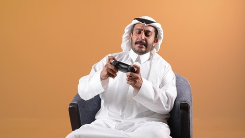 الحماس والتفاعل والاندماج مع اللعب، الاستمتاع بألعاب الفيديو المسلية، الاستفادة من التقنيات الحديثة والتكنولوجيا في أوقات الفراغ، تصوير لشاب عربي خليجي قطري يرتدي الثوب التقليدي والشماغ يجلس على كرسي ويمسك القبضة بيديه