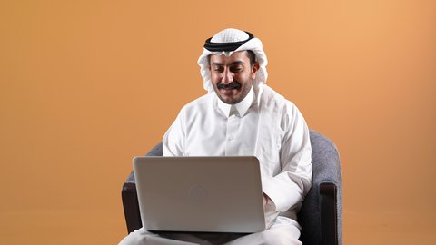 إقامة اجتماعات العمل عن بعد عبر الانترنت، إجراء مكالمات الفيديو باستخدام الحاسوب المحمول، مفهوم التوظيف أونلاين, دمج التقنية بالأعمال الإدارية، تصوير لرجل عربي خليجي قطري يرتدي الثوب التقليدي والشماغ يجلس على كرسي