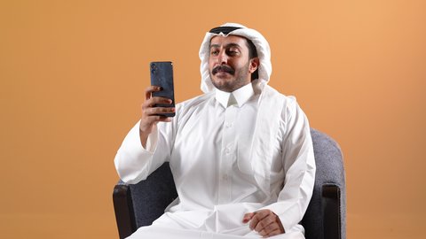 اجراء بث مباشر، دمج التقنية بالأعمال الإدارية، إجراء مكالمات الفيديو باستخدام الهاتف المحمول، إقامة اجتماعات العمل عن بعد, تصوير لرجل عربي خليجي قطري يرتدي الثوب التقليدي والشماغ يجلس على كرسي