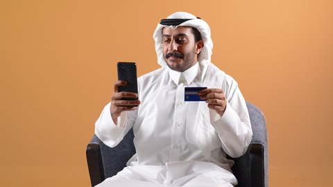 الدفع عن طريق بطاقه الائتمان ، التسوق عبر الانترنت ، دمج التقنية بالأعمال الإدارية، إجراء مكالمات الفيديو باستخدام الهاتف المحمول، إقامة اجتماعات العمل عن بعد, تصوير لرجل عربي خليجي قطري يرتدي الثوب التقليدي والشماغ يجلس على كرسي