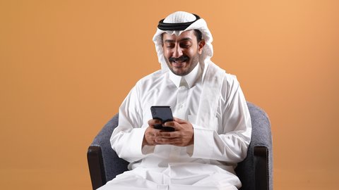 الدردشه عبر الانترنت ، اجراء مكالمه هاتفيه ، دمج التقنية بالأعمال الإدارية، إجراء مكالمات الفيديو باستخدام الهاتف المحمول، إقامة اجتماعات العمل عن بعد, تصوير لرجل عربي خليجي قطري يرتدي الثوب التقليدي والشماغ يجلس على كرسي