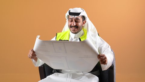 العمل في القطاع الهندسي، إتقان إدارة المشاريع باستخدام الحاسوب المحمول، مفهوم الهندسة والانشاءات المعماريه، تصوير مقرب من الجانب لمهندس عربي خليجي قطري يرتدي الثوب التقليدي والغتره مع سترة الحماية يجلس خلف طاولة المكتب وأمامه المخطط وبجانبه الخوذة