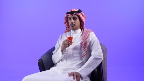 الحصول على عصير صيفي بارد ومنعش،إيماءات المفاجأة والاندماج الاستمتاع بمشاهدة الأفلام، تصوير لشاب عربي خليجي بحريني يرتدي الثوب التقليدي والشماغ يجلس على كرسي ويمسك بيده كأس من المشروب المفضل