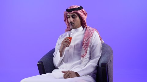 إيماءات المفاجأة والاندماج الاستمتاع بمشاهدة الأفلام، الحصول على عصير صيفي بارد ومنعش، تصوير لشاب عربي خليجي بحريني يرتدي الثوب التقليدي والشماغ يجلس على كرسي ويمسك بيده كأس من المشروب المفضل