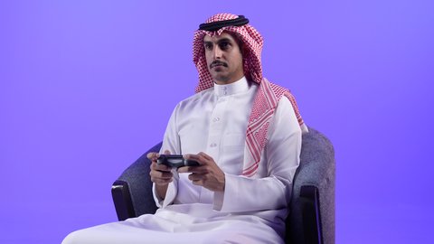 الحماس والتفاعل والاندماج مع اللعب، الاستمتاع بألعاب الفيديو المسلية، الاستفادة من التقنيات الحديثة والتكنولوجيا في أوقات الفراغ، تصوير لشاب عربي خليجي بحريني يرتدي الثوب التقليدي والشماغ يجلس على كرسي ويمسك القبضة بيديه