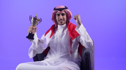 تشجيع الفرق والمنتخبات والاندية ، حب الوطن والانتماء اليه، رموز وثقافات وطنية،تصوير لشاب عربي خليجي بحريني يرتدي الثوب التقليدي والشماغ وعلم مملكة البحرين يجلس على كرسي مريح يحمل بيده كأس البطولة وكرة القدم ينظر الى مكان ما في الاعلى بإيماءات الاعتزاز والفخر والسرور