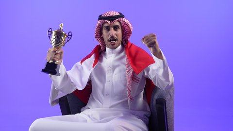 حب الوطن والانتماء اليه، رموز وثقافات وطنية، تشجيع الفرق والمنتخبات والاندية ، تصوير لشاب عربي خليجي بحريني يرتدي الثوب التقليدي والشماغ وعلم مملكة البحرين يجلس على كرسي مريح يحمل بيده كأس البطولة وكرة القدم ينظر الى مكان ما في الاعلى بإيماءات الاعتزاز والفخر والسرور
