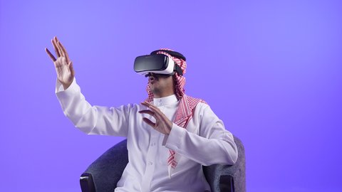 الاندماج والتفاعل ومحاكاة العالم الخيالي، تصوير لرجل عربي خليجي بحريني يرتدي الثوب التقليدي والشماغ ونظارات الواقع الافتراضي يجلس على كرسي مريح يقوم بمد يديه الى الامام بتعابير صد شيء ما والدفاع عن النفس، شاب سعودي يستخدم نظارة الفي آر