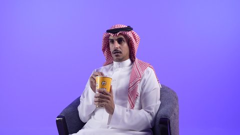 إيماءات الاندماج والتركيز مع الفيلم، الجلوس ومشاهدة الأفلام في أوقات الفراغ، الاستمتاع بتناول الفشار، تصوير لشاب عربي خليجي بحريني يرتدي الثوب التقليدي والشماغ يجلس على كرسي ويمسك بيده علبة من البوب كورن اللذيذ