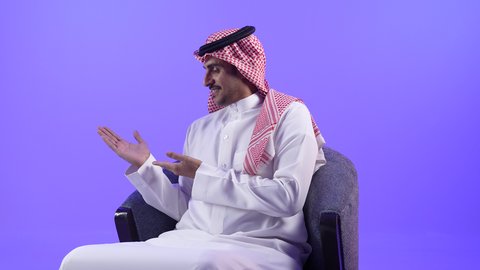 الاشارة والتحديد لشخص معين، شاب سعودي ينظر جانبا بإيماءات الفرح والسرور، تصوير لشاب عربي خليجي بحريني مبتسم يرتدي الثوب التقليدي والشماغ يجلس على كرسي مريح يشير بإصبعه السبابة إلى شيء محدد بتعابير هذا المكان هنا