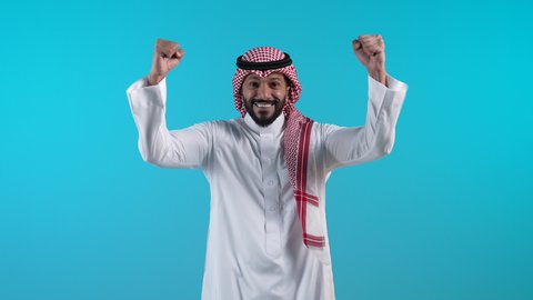 الانجاز وتحقيق الاهداف، تصويرلرجل عربي خليجي بحريني مبتسم يرتدي الثوب التقليدي والشماغ  يرفع قبضة يديه للاعلى بإيماءات الفوز والانتصار والنجاح, شاب سعودي ينظر الى الكاميرا بتعابير السعادة والقوة والسرور
