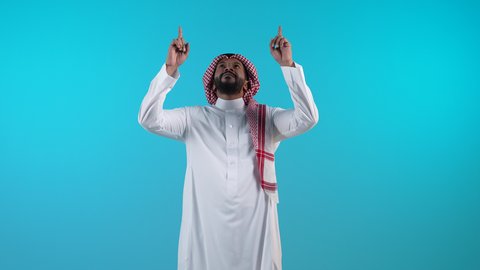 النظر الى الكاميرا بايماءات السعادة والسرور، تحديد موقع معين، الإشارة والتوضيح إلى شيء ما، تصوير لرجل عربي خليجي بحريني يرتدي الثوب التقليدي والشماغ يرفع اصبعه السبابة إلى الأعلى، خلفية زرقاء