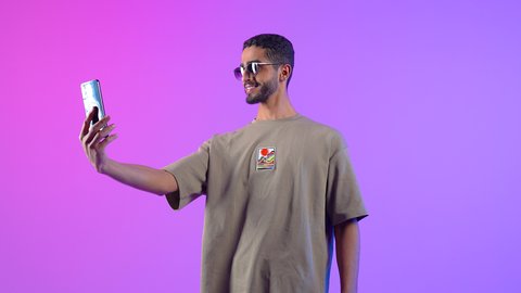 إجراء مكالمة فيديو بتعابير الفرح والسرور, دردشة و اتصالات مع الأهل والأصدقاء, بورتريه لشاب عربي خليجي عماني مبتسم يرتدي الزي الكاجوال والنظارة الشمسية يمسك بيده الهاتف و يقوم بالتقاط صورة سيلفي، خلفية ملونه