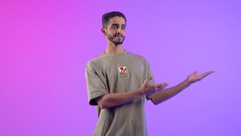 حمل عنصر هولوجرامي، تحديد موقع شيء ما، بورتريه لشاب عربي خليجي عمانى مبتسم يرتدي الزي الكاجوال يشير الى كفه بايماءات السرور والسعادة، مفهوم التقديم والعرض، رجل سعودي ينظر الى الكاميرا بتعابير البهجة،، خلفية ملونه