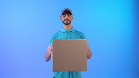 Safe delivery service, portrait of an Arab Gulf Omani delivery representative wearing casual clothes and a gray hat, smiling, holding cardboard boxes in his hand, delivering express orders, a Saudi man looking at the camera with gestures of pleasure, blue background