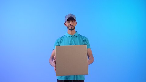 مفهوم التسوق الإلكتروني الآمن, بورتريه لمندوب توصيل عربي خليجي عماني يرتدي الزي الكاجوال و القبعه الرمادية مبتسم يحمل بيده علب كرتونية يقوم بتوصيل الطلبات السريعه، رجل سعودي ينظر إلى الكاميرا بإيماءات السرور، خلفية زرقاء