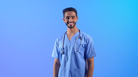 الوقوف باستعداد والنظر إلى الكاميرا بإيماءات السرور، الاستعداد لتقديم الخدمات الطبية للمرضى, مفهوم الطب والرعاية الصحية، ممرض سعودي يعمل بالقطاع الطبي, بورتريه لشاب عربي خليجي عماني مبتسم يرتدي يونيفورم و السماعات الطبيه العمل يقف باستقامة، خلفية زرقاء