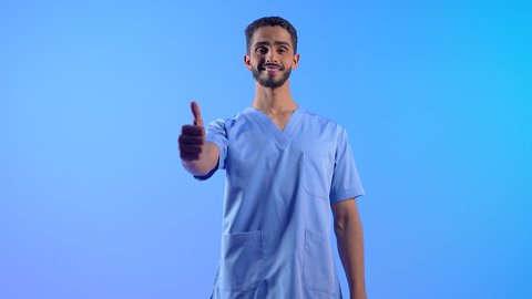 Quality and excellence mark, concept of medicine and healthcare, readiness to provide medical services to patients, Saudi nurse working in the medical sector, raising his thumb up in a gesture of acceptance, portrait of a smiling young Arab Gulf Omani man wearing a work uniform standing straight, blue background