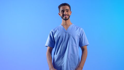 الاستعداد لتقديم الخدمات الطبية للمرضى, مفهوم الطب والرعاية الصحية، ممرض سعودي يعمل بالقطاع الطبي، الوقوف باستعداد والنظر إلى الكاميرا بإيماءات السرور, بورتريه لشاب عربي خليجي عماني مبتسم يرتدي يونيفورم العمل يقف باستقامة