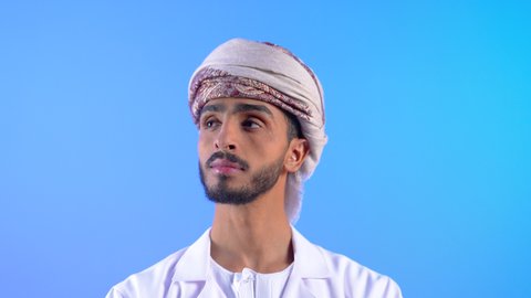 مفهوم الرجولة والوسامة، الوقوف بأيدي متقاطعة بإيماءات الثقة بالنفس، بورتريه لشاب عربي خليجي عماني يرتدي الدشداشه و العمامة ينظر إلى مكان ما بتعابير السرور والاندماج، الاهتمام بالمظهر الخارجي والأناقة، خلفية زرقاء