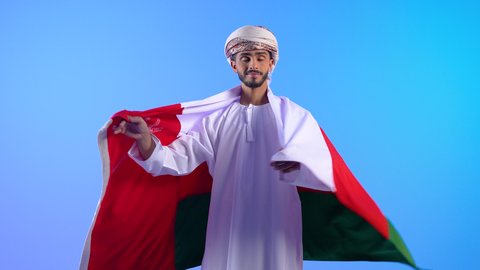 الفخر بالرموز والراية العمانية، الاعتزاز بالانتماء للبلاد، الاحتفال بذكرى اليوم الوطني، بورتريه لرجل عربي خليجي عماني يرتدي الدشداشة والكمه يضع علم سلطنة عمان على ظهره وينظر للكاميرا بإيماءات السرور والسعادة، مفهوم حب الوطن، خلفية زرقاء