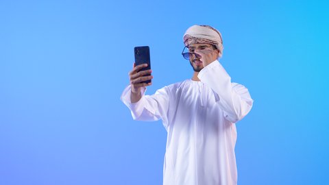 إلتقاط صورة سيلفي تذكارية بالهاتف المحمول، بورتريه لشاب عربي خليجي اماراتي يرتدي الدشداشة و العمامه مبتسم يرتدي نظارة شمسية ، إجراء مكالمة فيديو ، صورة للذكرى ، خلفية زرقاء