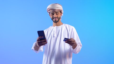 إمكانية تسديد الفواتير عبر بطاقة الصراف, الشراء والدفع عن طريق بطاقات الإئتمان البنكية، بورتريه لشاب عربي خليجي عماني مبتسم يرتدي االدشداشة و العمامة  يمسك بيده البطاقة الالكترونية و الهاتف المحمول بايماءات السعادة و السرور , خلفية زرقاء