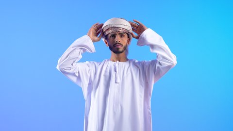 الاهتمام بالأناقة والمظهر الخارجي، مفهوم القوة والرجولة، بورتريه لرجل عربي خليجي عماني يرتدي الدشداشه والعمامة ينظر الى الكاميرا بتعابير السعادة و السرور، ايماءات الثقة بالنفس، خلفية زرقاء
