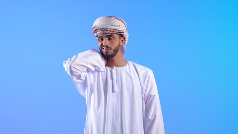 آلام المفاصل والعظام والعمود الفقري، تعابير الوجه تدل على الاضطراب والتألم من الكتفين، بورتريه لرجل عربي خليجي عماني يرتدي الدشداشة و العمامة يمسك كتفه بإيماءات الألم, الحاجة للراحة والحصول على العلاج، خلفية زرقاء