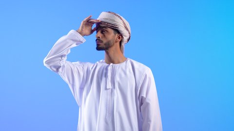 تعابير محاولة رؤية الأشياء البعيدة, مفهوم التطلع للمستقبل, النظر لمكان ما, بورتريه لرجل عربي خليجي عماني يرتدي الدشداشة و العمامة  يرفع يده إلى رأسه بإيماءات البحث عن شيء محدد, خلفية زرقاء