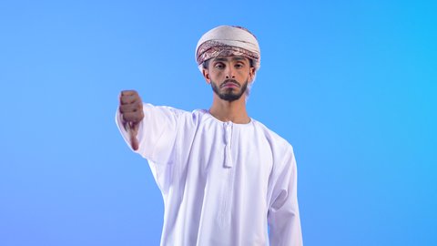 مفهوم الرفض وعدم القبول، بورتريه لشاب عربي خليجي عماني يرتدي الدشداشة و العمامة يشير بإصبع الابهام للاسفل بإيماءات عدم الاعجاب والتقييم السلبي، الشعور بالانزعاج من شيء ما، خلفية زرقاء
