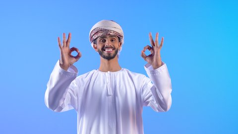 Expressing admiration and appreciation, raising hands with signs of quality and acceptance, excellence and full marks, feeling satisfied and appreciative, portrait of a smiling young Arab Gulf Omani man wearing a dishdasha and turban looking at the camera with gestures of happiness and pleasure, blue background