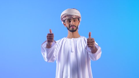 ايماءات الإعجاب والموافقة، علامة الجودة والامتياز، الوقوف باستقامة بتعابير الثقة بالنفس، بورتريه لشاب عربي خليجي عماني مبتسم يرتدي الدشداشة و العمامة ينظر إلى الكاميرا بايماءات السرور والبشاشة، خلفية زرقاء