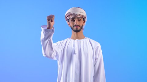 علامة الجودة والامتياز، ايماءات الإعجاب والموافقة،  الوقوف باستقامة بتعابير الثقة بالنفس، بورتريه لشاب عربي خليجي عماني مبتسم يرتدي الدشداشة و العمامة ينظر إلى الكاميرا بايماءات السرور والبشاشة، خلفية زرقاء