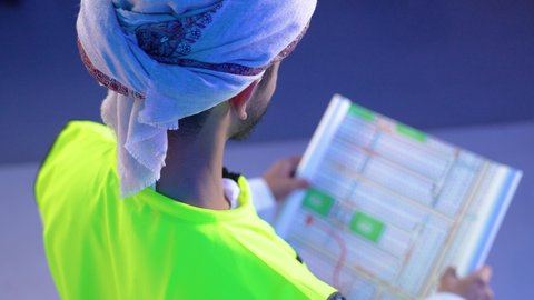 العمل في القطاع الهندسي، مفهوم الهندسة والانشاءات المعمارية، بورتريه مقرب من الخلف لمهندس عربي خليجي عماني يرتدي الدشداشة و العمامة وسترة الحماية يمسك المخطط ينظر إليه بايماءات التركيز والتمعن، مهنة ووظيفة للشباب في سلطنة عمان، خلفية زرقاء