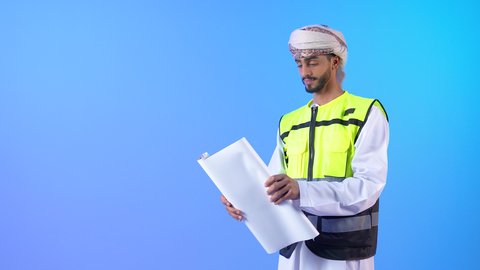 مفهوم الهندسة والانشاءات المعمارية، بورتريه لمهندس عربي خليجي عماني يرتدي الدشداشة و العمامة وسترة الحماية يمسك المخطط ينظر إليه بايماءات التركيز والتمعن، مهنة ووظيفة للشباب في سلطنة عمان، العمل في القطاع الهندسي، خلفية زرقاء