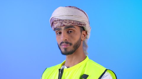 مفهوم الهندسة والانشاءات المعمارية، بورتريه مقرب لمهندس عربي خليجي عماني مبتسم يرتدي العمامة وسترة الحماية  ينظر إلى الكاميرا بإيماءات السرور، الوقوف بأيدي متقاطعه، العمل في القطاع الهندسي، مهن ووظائف سلطنه عمان، الإعمار بسواعد أبناء الوطن، خلفية زرقاء