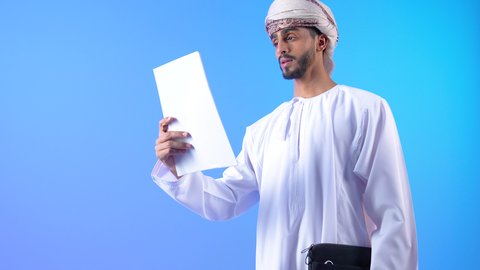 Preparing reports and work tasks, the concept of work, diligence and completion of tasks, a portrait of a young Arab Gulf Omani man wearing a dishdasha and turban, holding a bag and work papers in his hand, blue background