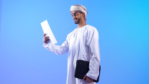The concept of business and project management, going to work actively, a portrait of a young Arab Gulf Omani man wearing a dishdasha and turban, carrying a bag in his hand and reading work papers, taking care of his outward appearance, blue background