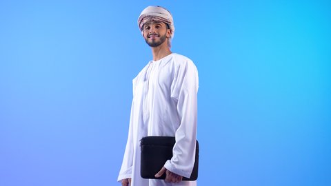 الذهاب إلى العمل بنشاط، الوقوف باستقامة والنظر إلى الكاميرا، بورتريه لشاب عربي خليجي عماني يرتدي الدشداشة و العمامه يحمل بيده حقيبة ، الاهتمام بالمظهر الخارجي، مفهوم إدراة الأعمال والمشاريع، خلفيه زرقاء