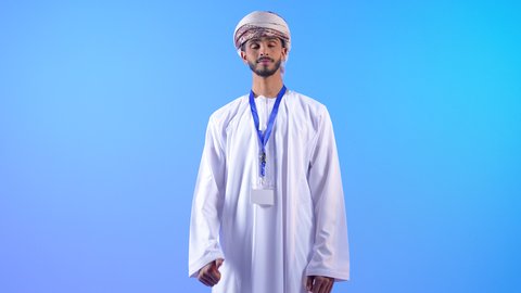 مفهوم الرجولة والأناقة، الوقوف باستقامة والنظر إلى الكاميرا، بورتريه لرجل عربي خليجي عماني يرتدي الدشداشة و العمامة يضع الهوية الشخصية حول رقبته ينظر بإيماءات الجدية والثقة بالنفس، الاهتمام بالمظهر الخارجي, خلفية زرقاء