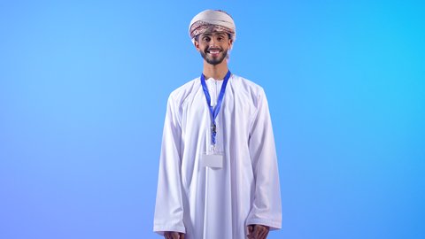 الوقوف باستقامة والنظر إلى الكاميرا، بورتريه لرجل عربي خليجي عماني يرتدي الدشداشة و العمامة يضع الهوية الشخصية حول رقبته ينظر بإيماءات الجدية والثقة بالنفس, مفهوم الرجولة والأناقة، الاهتمام بالمظهر الخارجي, خلفية زرقاء